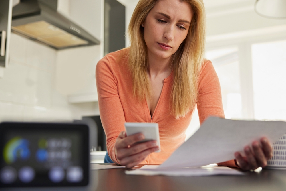 A photo of a woman looking worried at an energy bill. She is holding a phone and a smart meter can be seen in the forefront of the image.