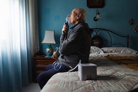 Photograph of an older man using a breathing apparatus, sitting on a bed. He is looking out of the window.