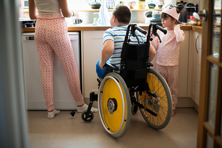 A woman at a kitchen sink with two children, a boy and a girl. The boy uses a wheelchair.