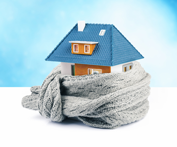 A toy house on a white surface behind a blue sky. The house is wrapped in a big woolly scarf.