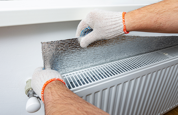 Photo of a person putting radiator reflector panels behind an electric radiator. You can only see the two hands of said person wearing gloves.