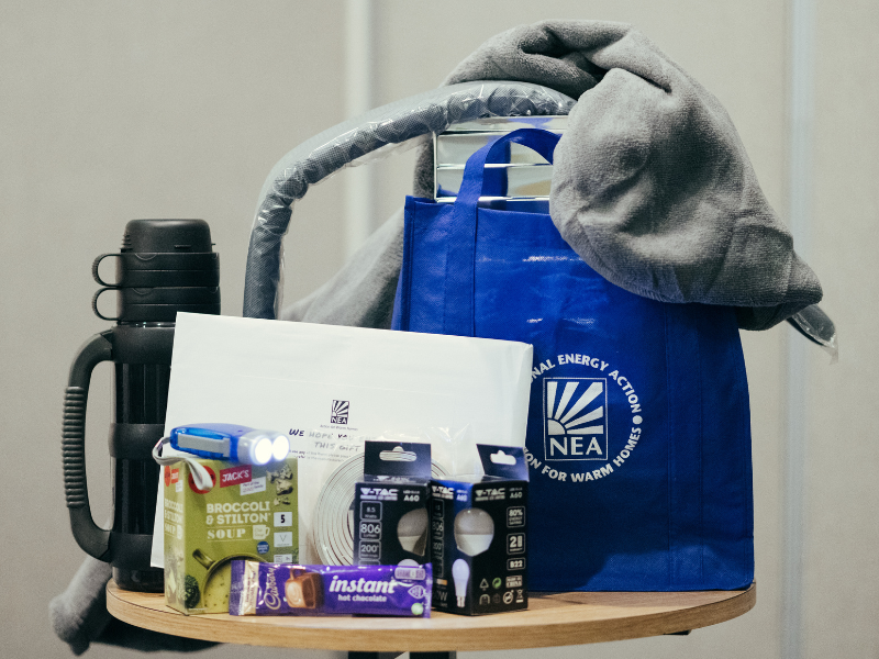 Photo of one of National Energy Action's Winter Warmth Support Packs. There is a blue bag with the logo on it, some energy efficient light bulbs, a thermos flask, hot chocolate. These are small measure that can be used to keep households in fuel poverty a little warmer.