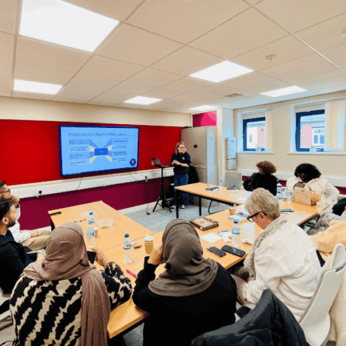 Seven adults at a community advice session. They are listening to an Adviser from National Energy Action giving a presentation on how to save money on their energy bills.