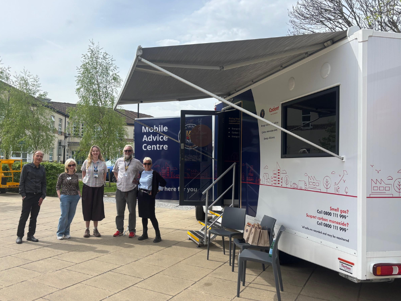A photo of five people stood next to National Energy Action's Mobile Advice Centre. The Advice Centre is a large van which is navy blue and white. There are two chairs underneath a covered area so we can provide energy advice to people regardless of poor weather.